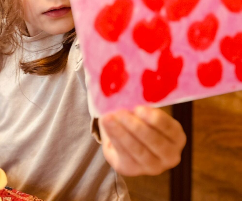 Carte postale décorée de gros cœurs rouges peints sur fond rose, tenue à la main par une enfant