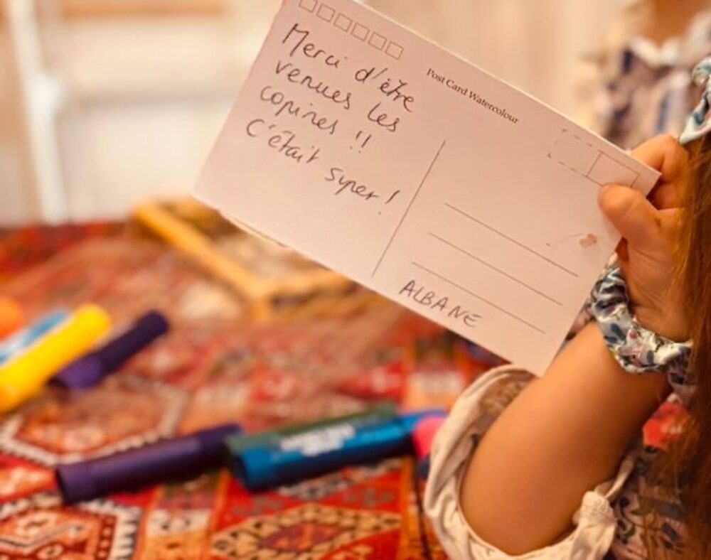 Carte postale vierge tenue à la main, avec un petit mot écrit au feutre sous la dictée d'un enfant : Merci d'être venues les copines, c'était super, signé Albane