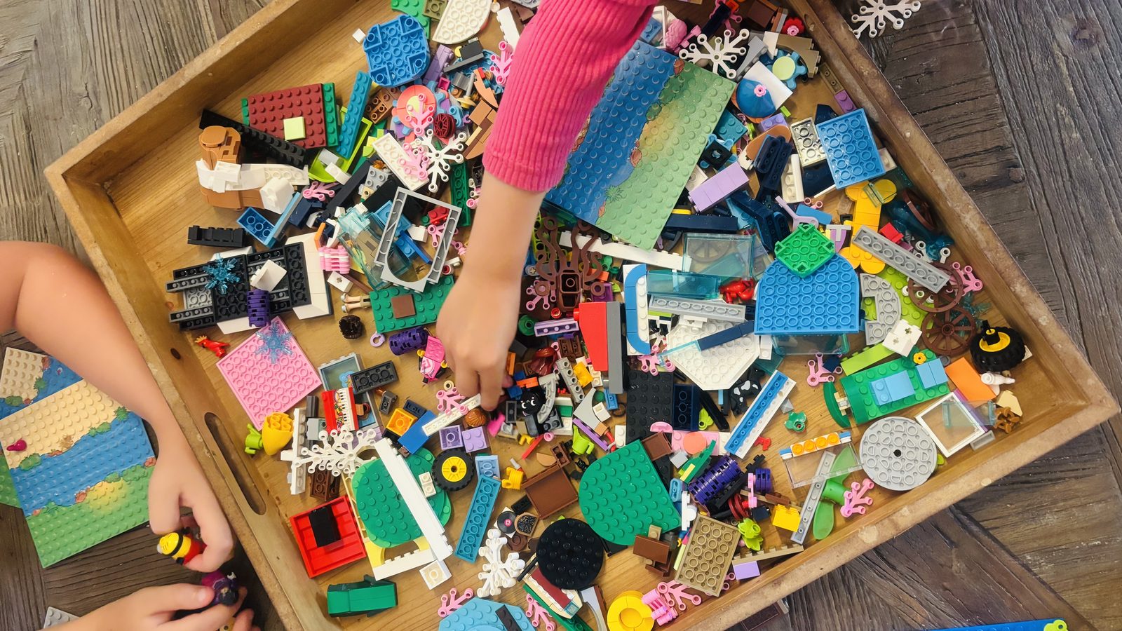 Deux enfants fouillent dans un grand plateau en bois rempli de briques Lego multicolores étalées en vrac, posé sur le sol