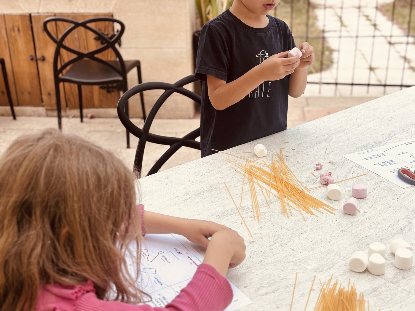 Deux enfants autour d'une table de jardin couverte de spaghettis crus et de chamallows, l'un tient une guimauve, l'autre suit du doigt une fiche imprimée