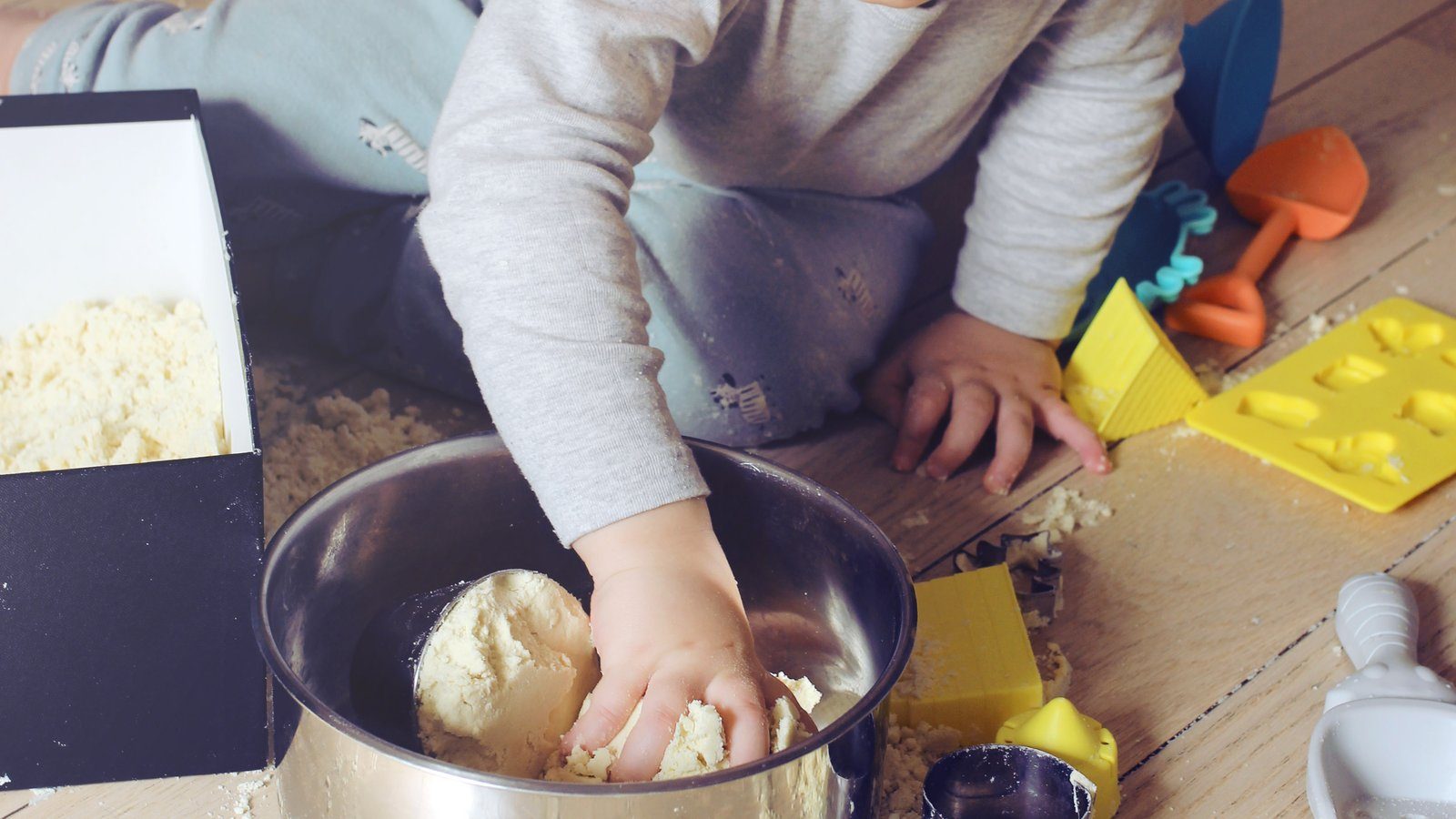 Un jeune enfant malaxe du sable magique maison dans un saladier en inox posé au sol, entouré de moules de plage