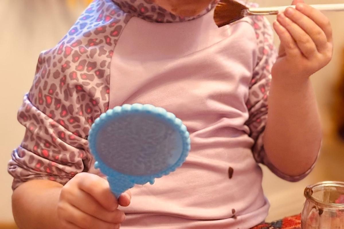Mains d'enfant tenant un pinceau chargé de chocolat fondu au-dessus d'un petit miroir bleu, un pot de chocolat posé sur la table, une tache de chocolat sur le tee-shirt
