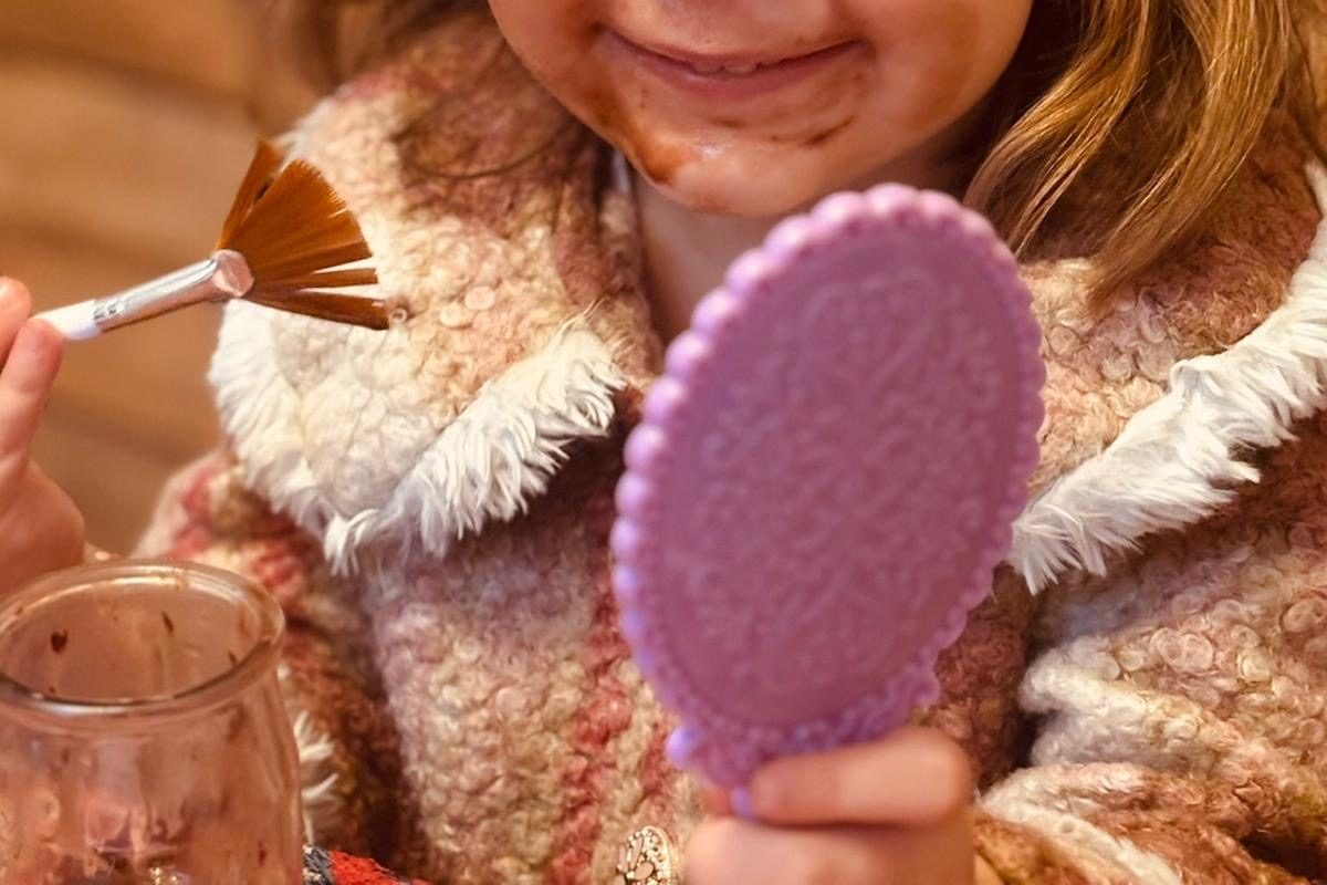 Enfant souriant, le bas du visage barbouillé de chocolat, qui s'admire dans un petit miroir violet en tenant un pinceau éventail, un pot de chocolat à côté
