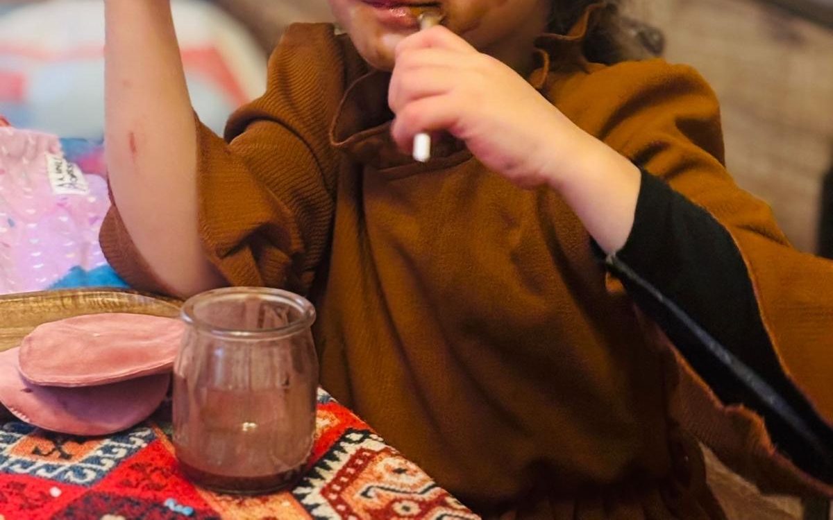 Enfant portant à sa bouche le manche d'un pinceau au chocolat, l'autre main levée tenant un miroir rose, un pot de chocolat sur la table