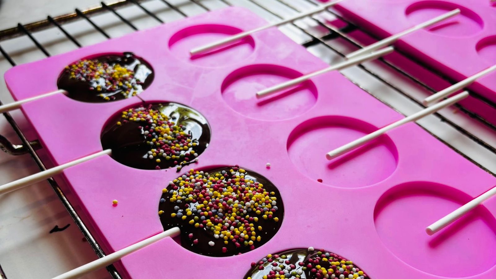 Gros plan sur un moule à sucettes rose posé sur une grille, disques de chocolat recouverts de vermicelles multicolores, bâtonnets blancs alignés