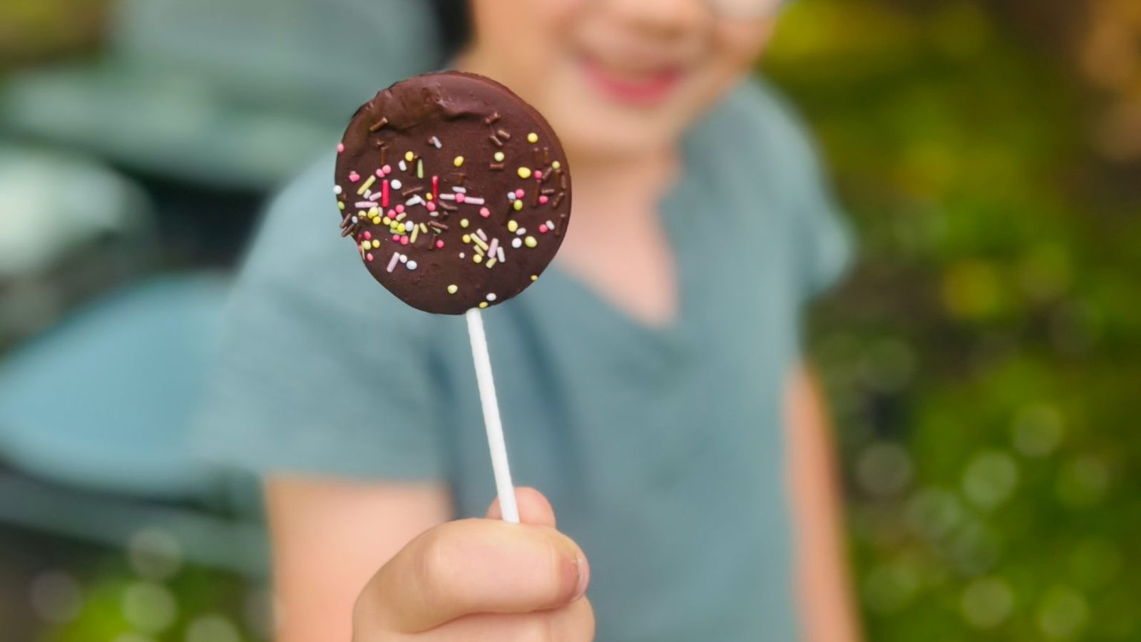 Main d'enfant tendant vers l'objectif une sucette en chocolat ronde décorée de vermicelles multicolores, jardin flou en arrière-plan
