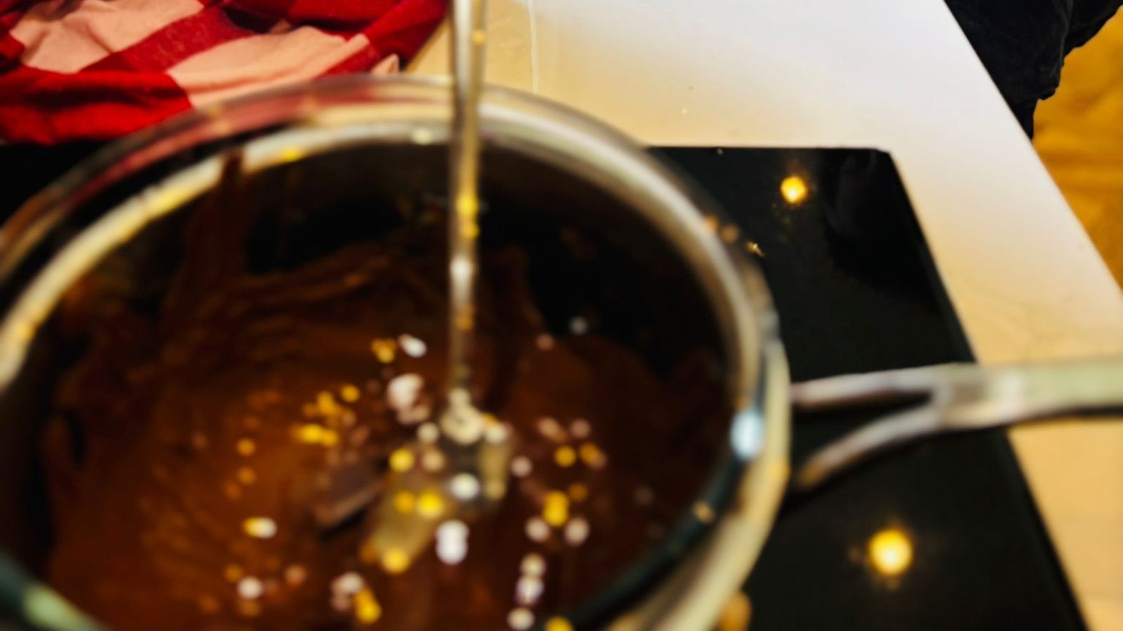 Casserole en inox remplie de chocolat noir fondu, lisse et brillant, avec un ustensile, quelques vermicelles tombés dedans, posée sur le plan de travail