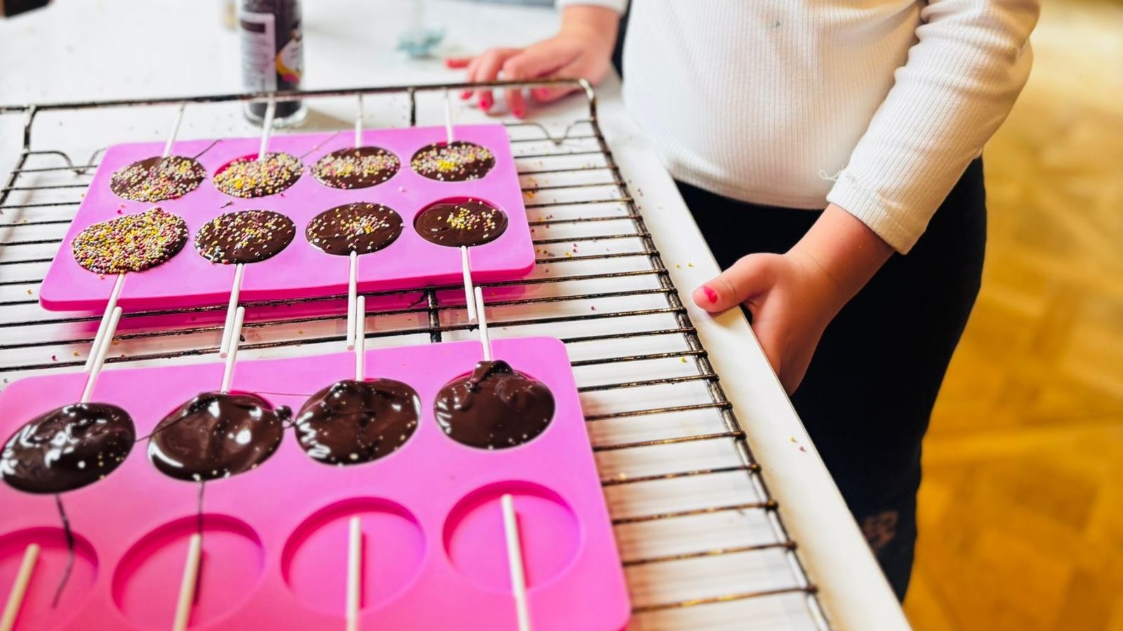 Deux moules à sucettes en silicone rose sur une grille, l'un déjà décoré de vermicelles colorés, l'autre rempli de chocolat brillant tout juste coulé avec les bâtonnets blancs en place