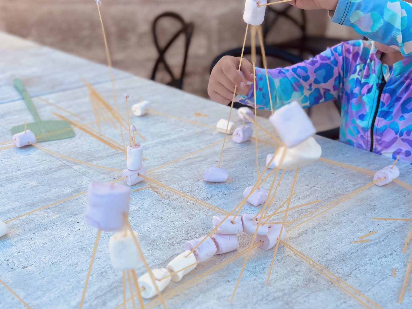 Une table de jardin couverte de spaghettis crus et de chamallows assemblés en petites structures, une enfant plante un spaghetti dans une guimauve