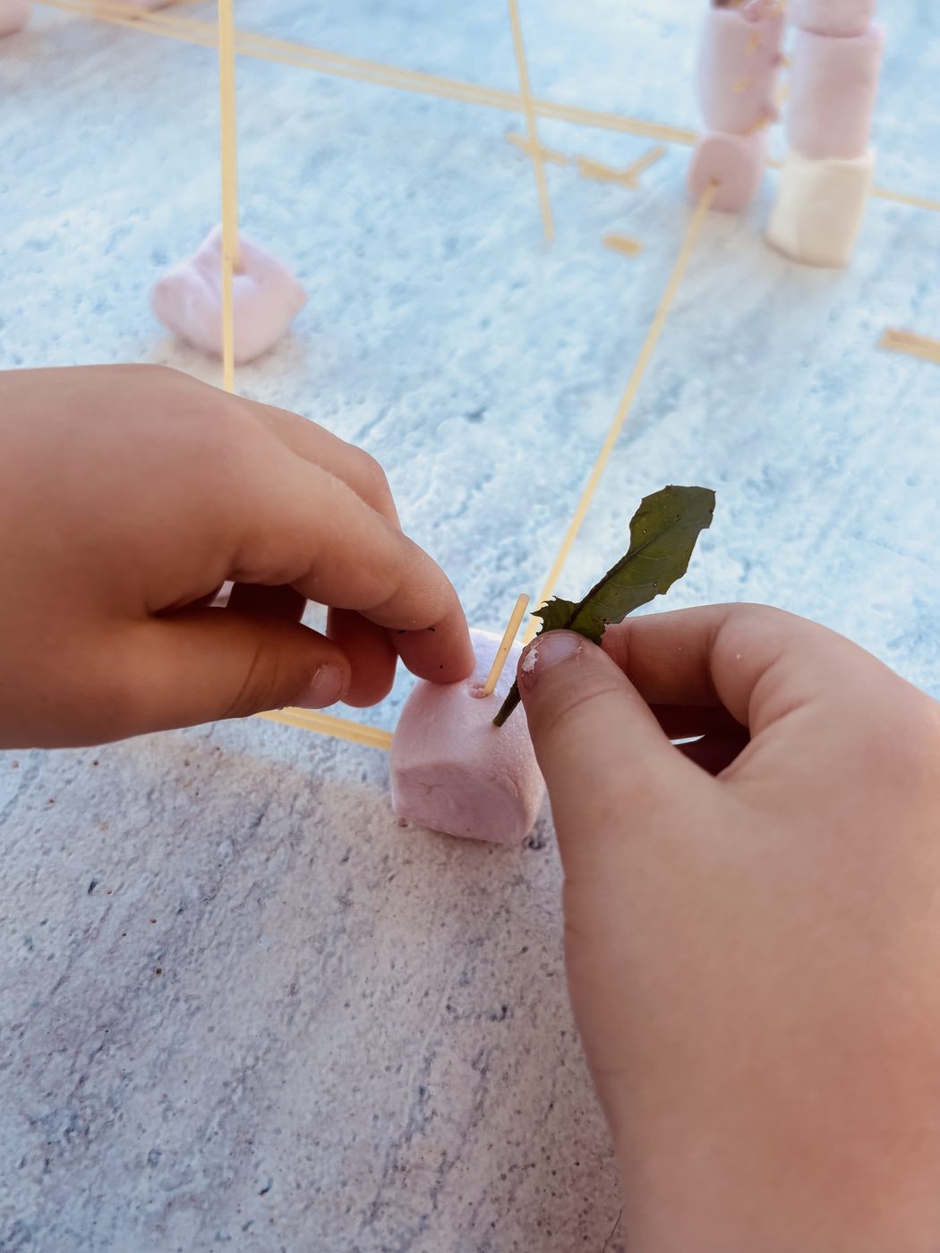 Deux mains d'enfant en gros plan enfonçant un spaghetti dans un chamallow rose posé sur la table