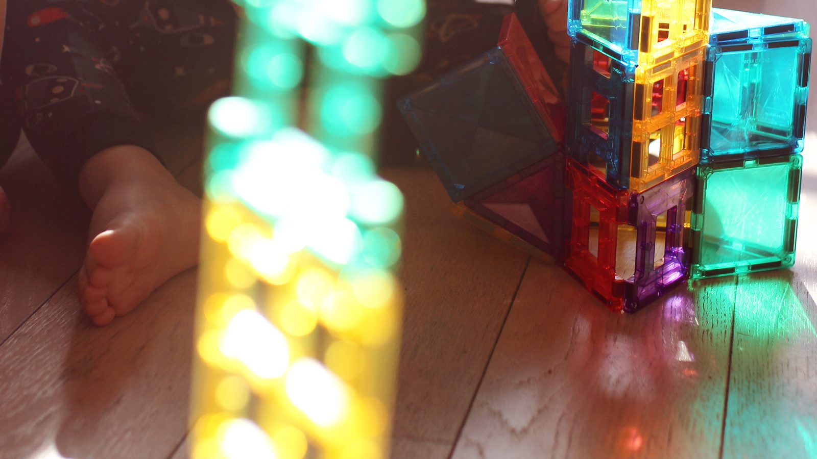 Tour de tuiles magnétiques traversée par le soleil au premier plan, construction de cubes colorés posée sur le parquet derrière