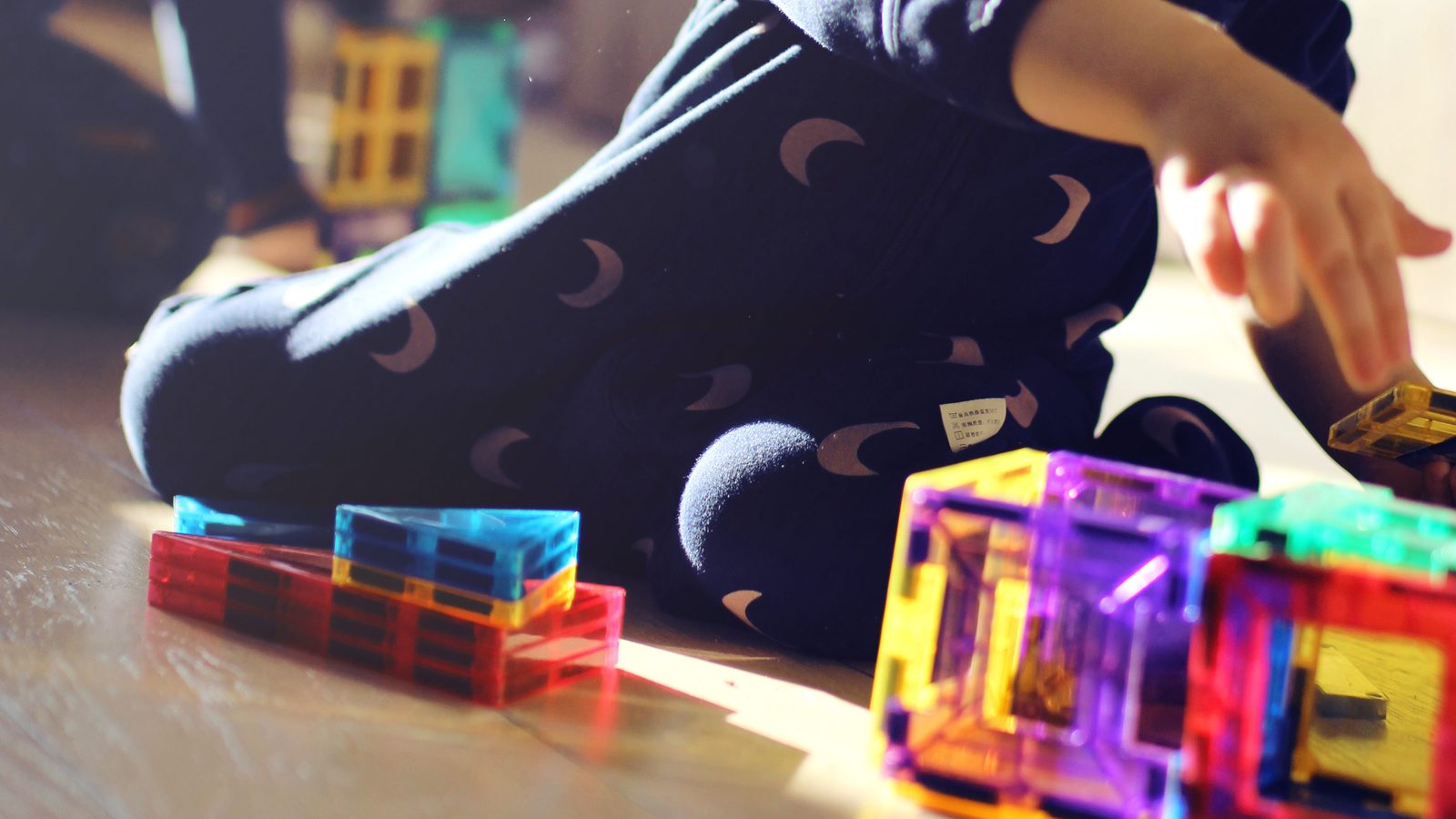 Main d'enfant attrapant une tuile magnétique jaune, cubes translucides multicolores posés sur le parquet au premier plan