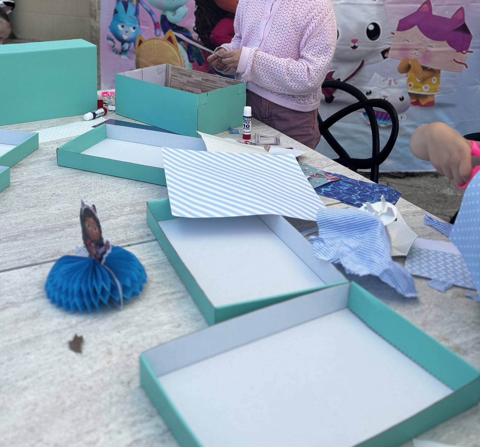 Atelier maison de poupée en cours : des boîtes turquoise à décorer sur la table, devant une toile Gabby et la Maison Magique
